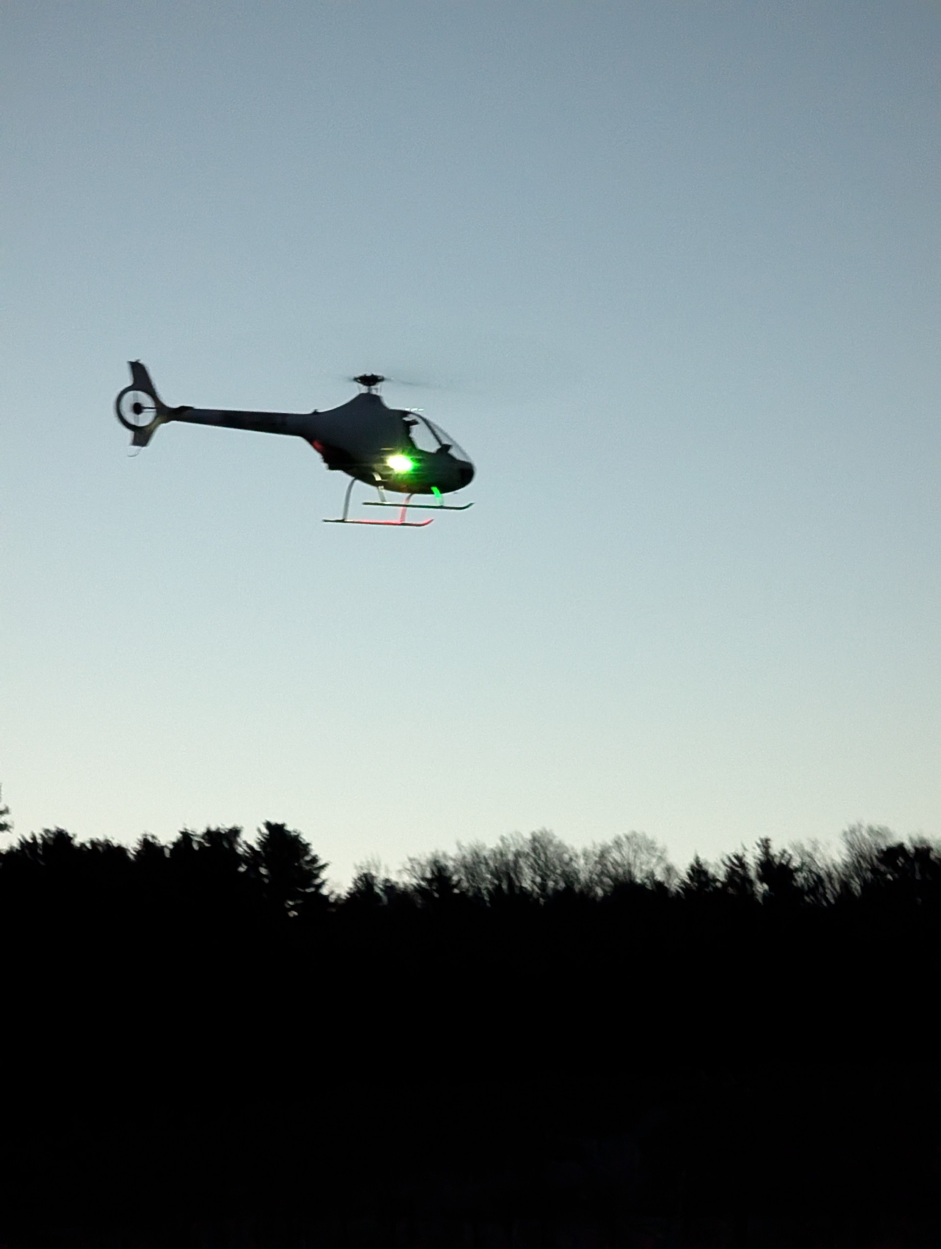 Helicopter with navigation lights flying low over a silhouetted treeline at dusk against a pale gray sky.